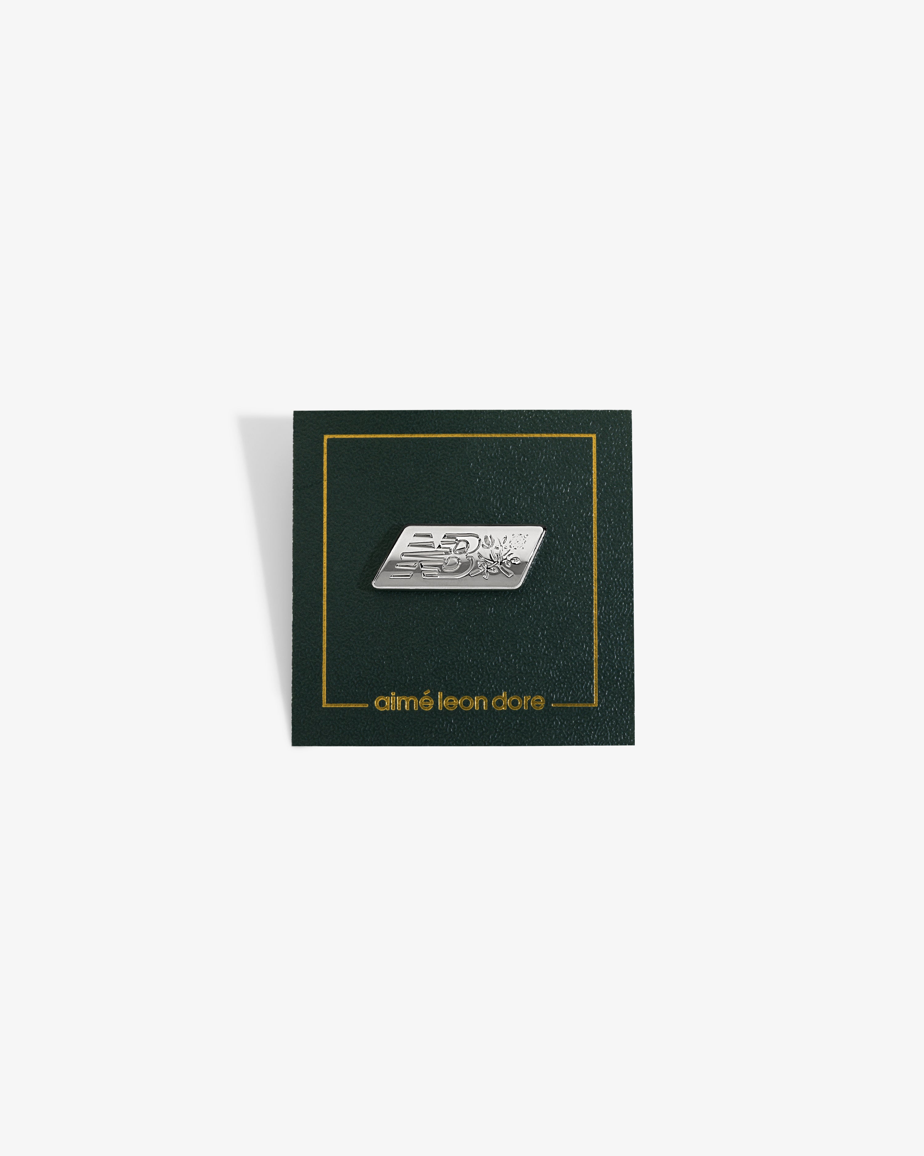 ALD / New Balance Logo Pin: silver NB on dark green card with gold border, aimé leon dore text at bottom.
