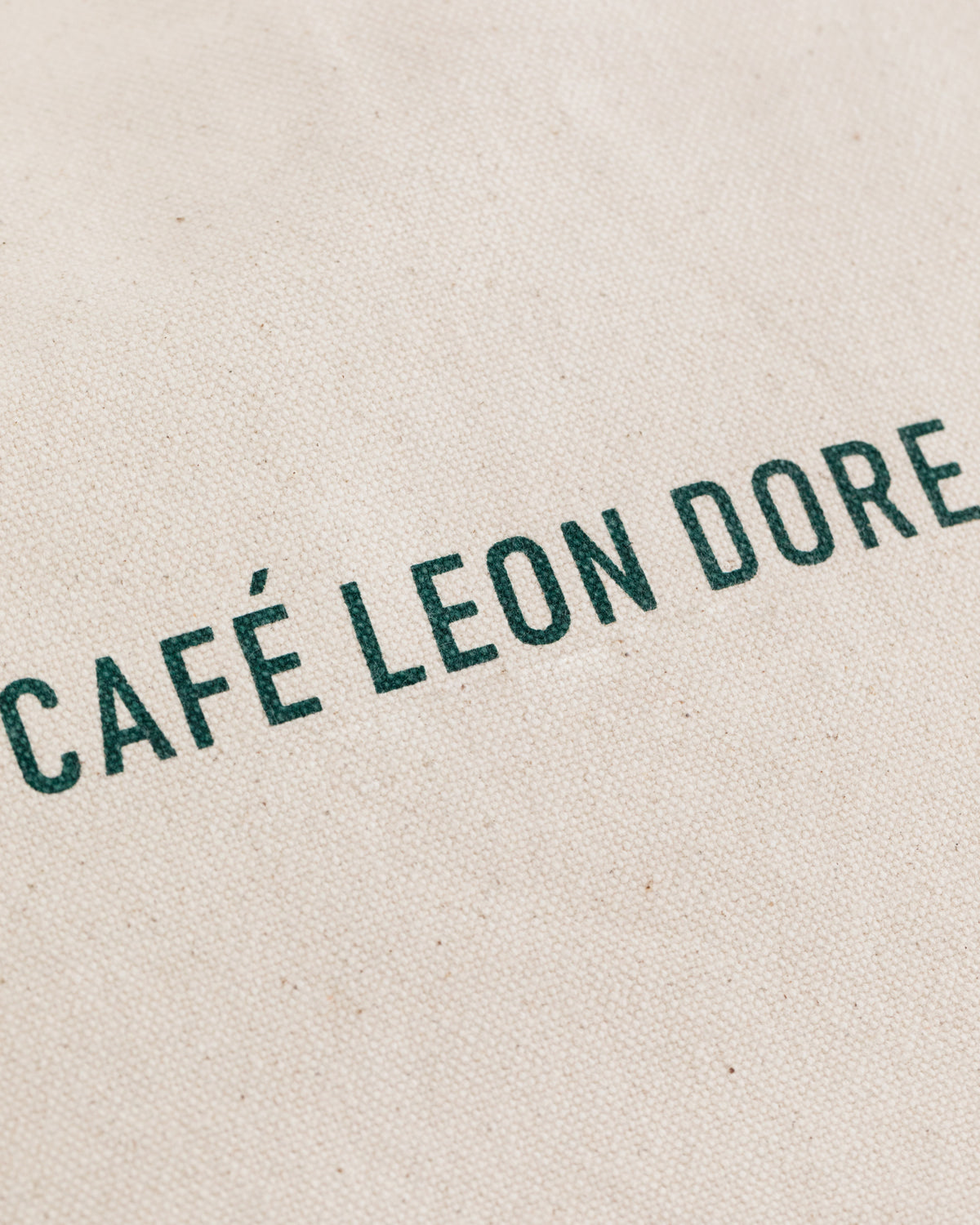 Close-up of Café Leon Dore Tote Bag made from beige fabric with green CAFÉ LEON DORE text.