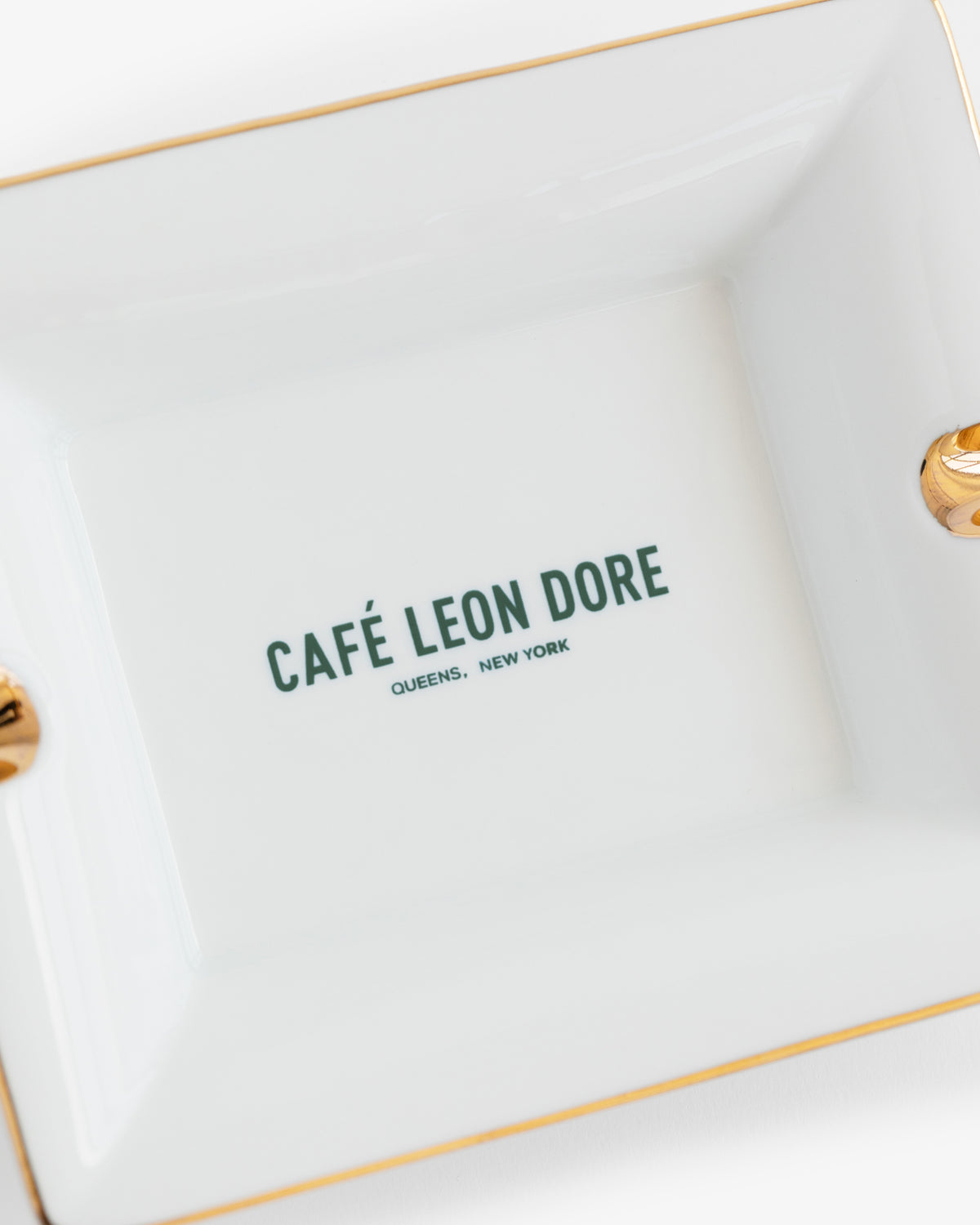 A white Café Leon Dore Ashtray with gold trim and "CAFÉ LEON DORE QUEENS, NEW YORK" printed in green at the center.