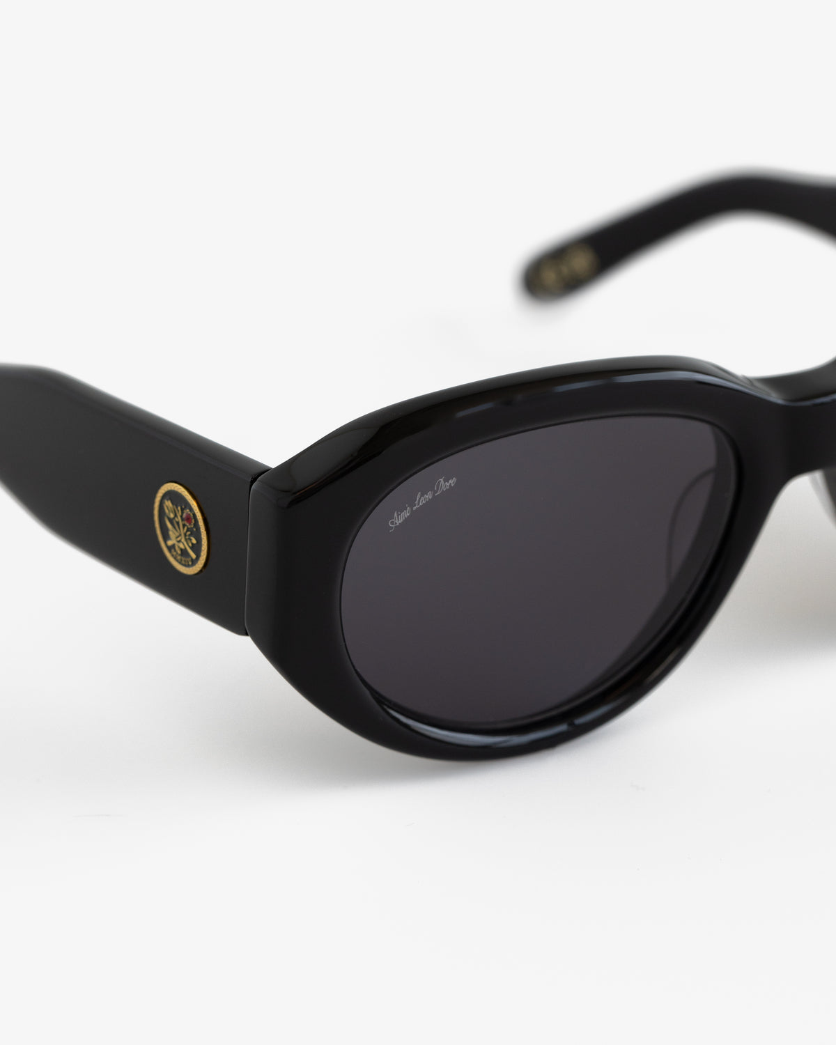 Jackie O Sunglasses: black oval frames, dark lenses, gold emblem on thick arms, shown on a white background.