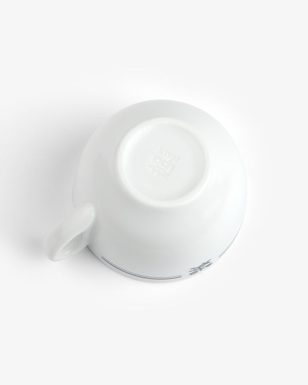A Café Leon Dore Cappuccino Cup is upside down on a white surface, revealing its base and part of the handle.