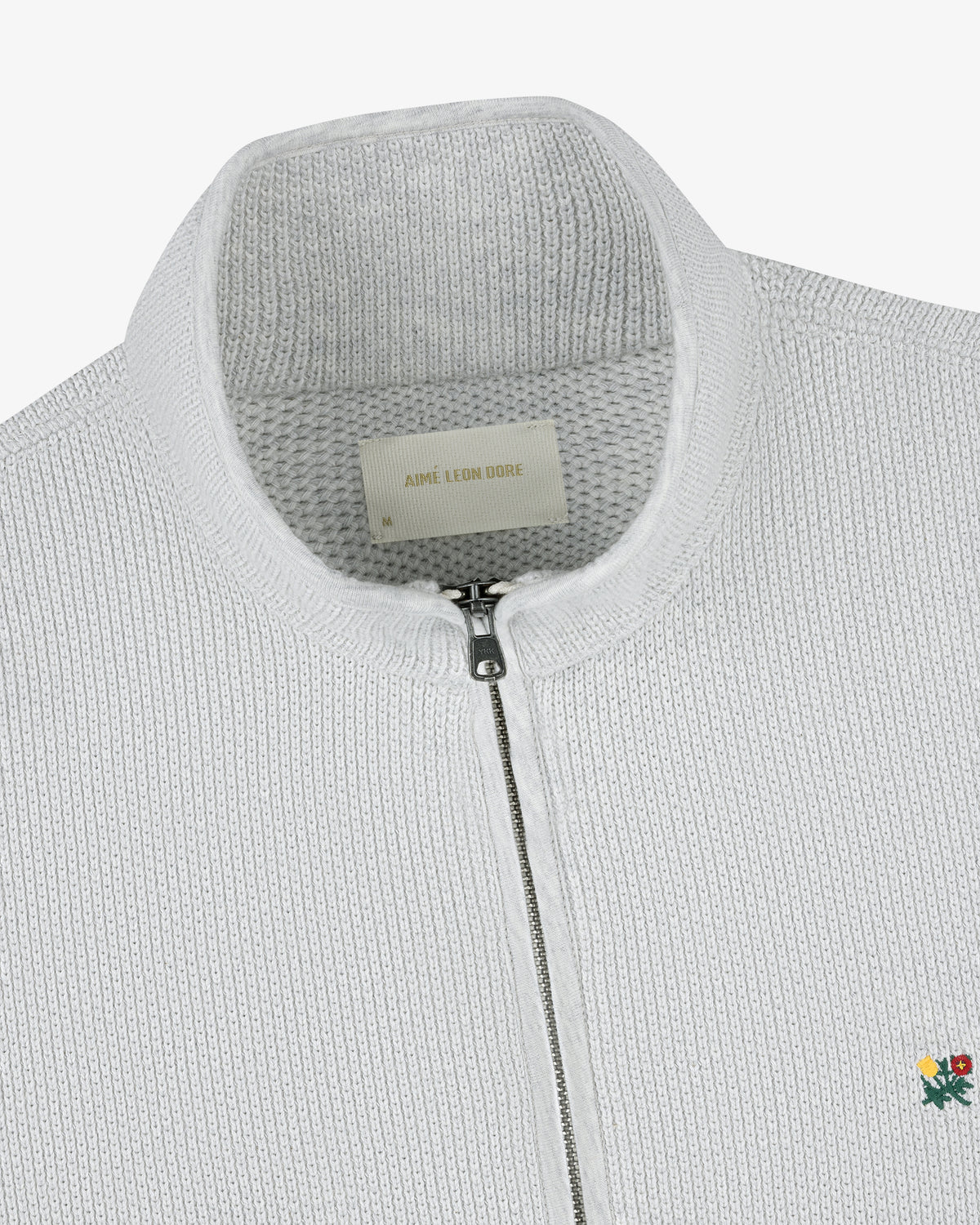 Close-up of the Crest Piqué Half-Zip Pullover, featuring a white knit fabric, embroidered flower, and Aimé Leon Dore label.