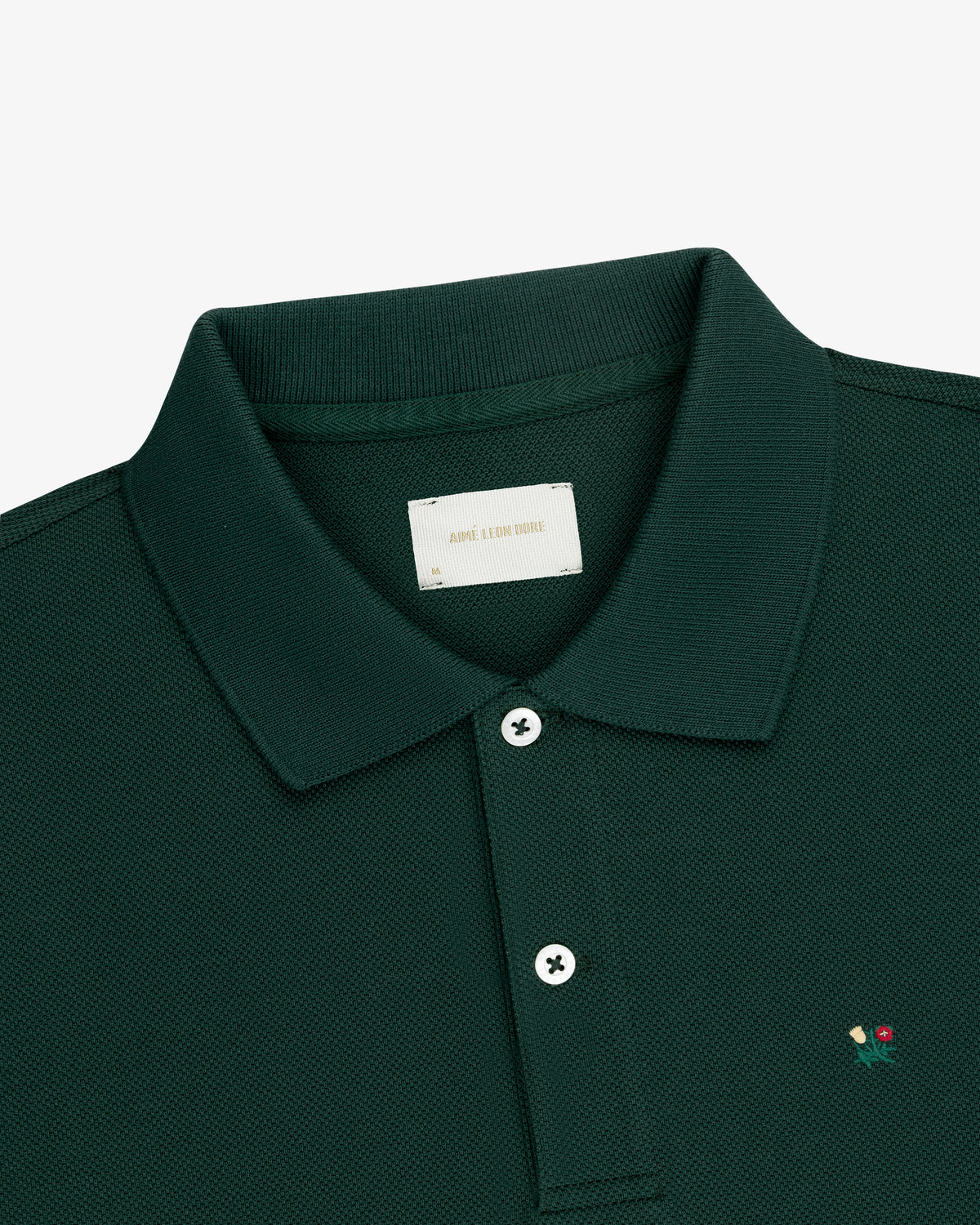 Crest Piqué Polo in dark green with two buttons and a small embroidered flower, viewed from the collar down.