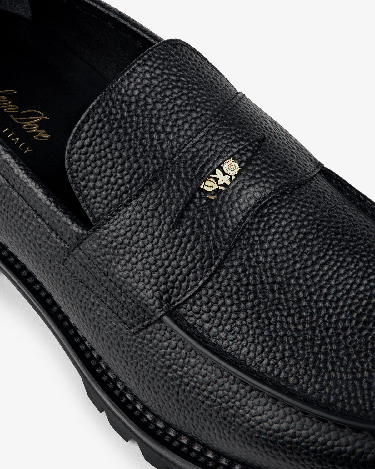 Close-up of the Lug Sole Queens Crest Loafer in black textured leather with a small gold emblem on the strap.