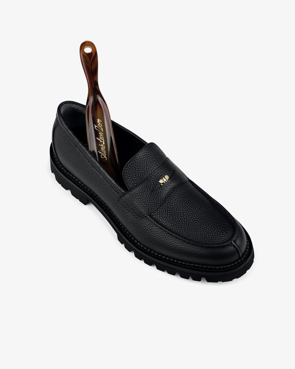 A Lug Sole Queens Crest Loafer in black leather with a gold logo and brown shoehorn, shown on a white background.
