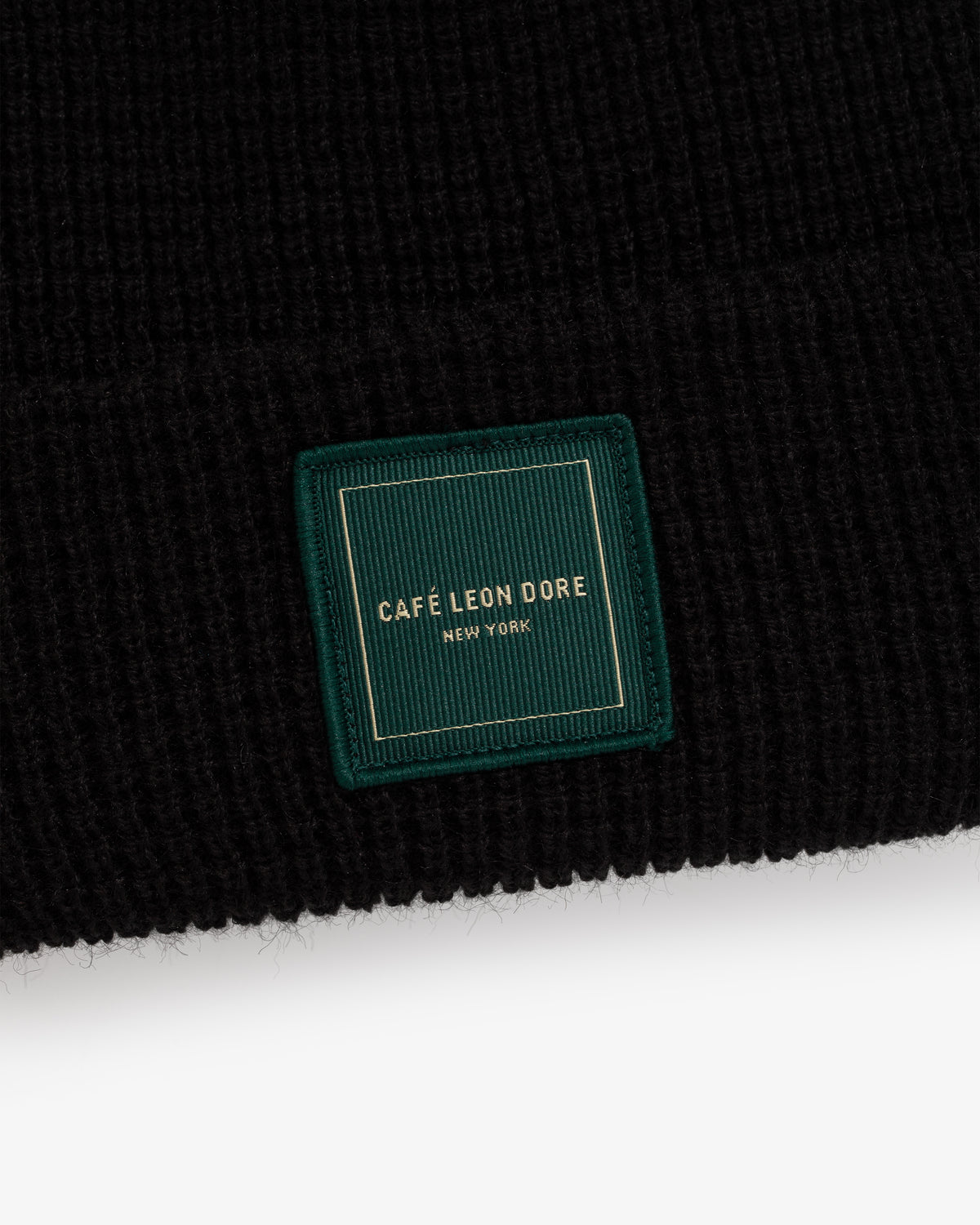 Close-up of a Café Leon Dore Beanie in black knit with a green patch label reading Café Leon Dore New York in gold.
