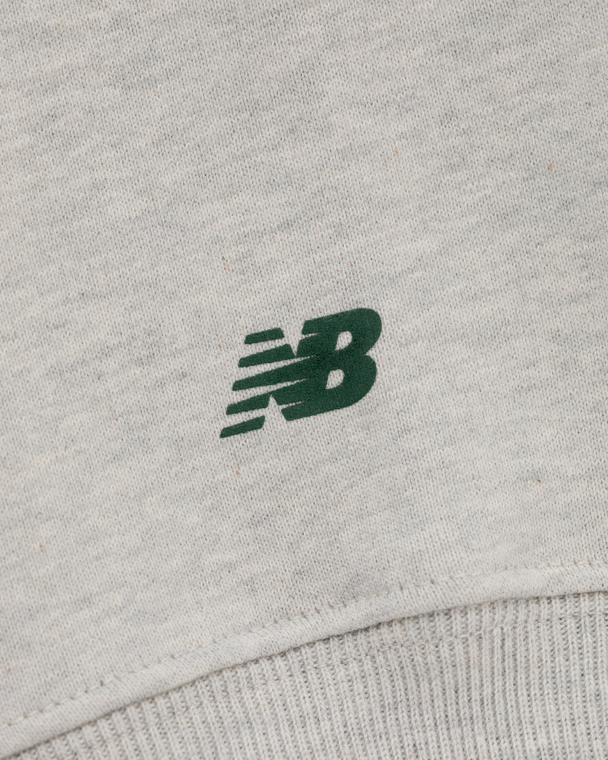 Close-up of ALD / New Balance SONNY NY IFTB Crewneck with dark green NB logo and ribbed hem on grey fabric.