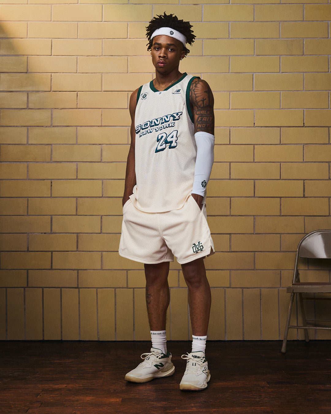 A basketball player in a white uniform and headband wears ALD / New Balance SONNY NY Calf Sleeves indoors by a brick wall.
