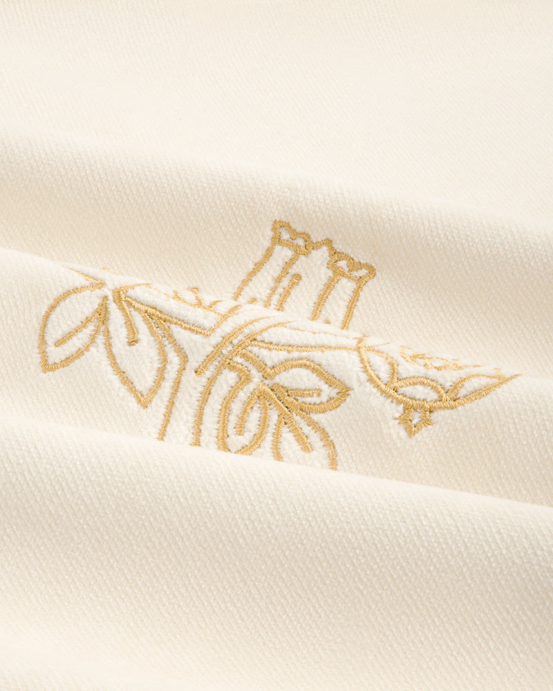 Close-up of Queens Crest Throw Pillow with cream fabric, gold floral embroidery, and visible textured folds.