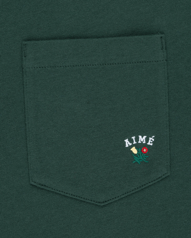 The Crest Pocket Tee is green with “AIMÉ” text and a small flower embroidered in yellow, green, and red.