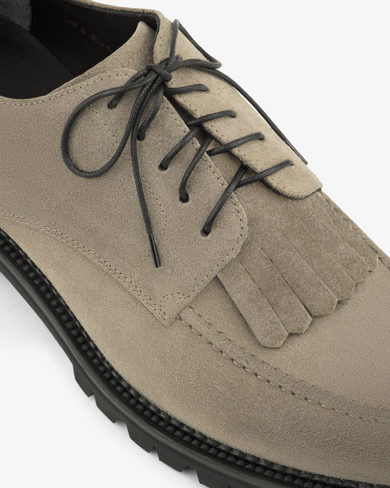 Close-up of the Suede Lug Sole Derby in beige with black laces, decorative fringe, and a textured black sole.