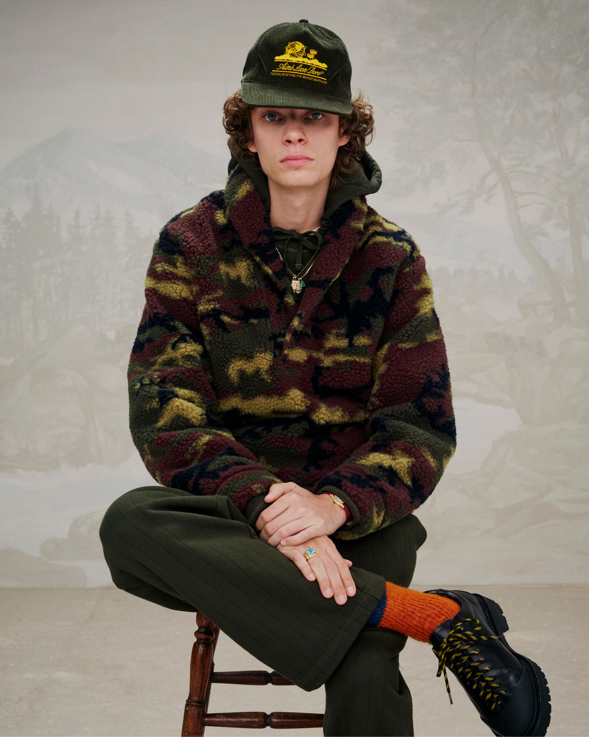 Wearing camo fleece, green pants, and a green hat, a person sits with hands folded in Leather Derby Hiker boots.