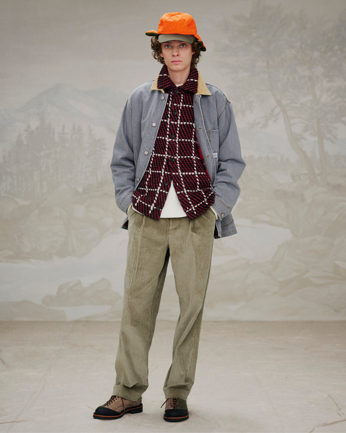 A person in autumn layers and orange cap stands indoors before a painted outdoor scene, wearing Suede Derby Hiker shoes.