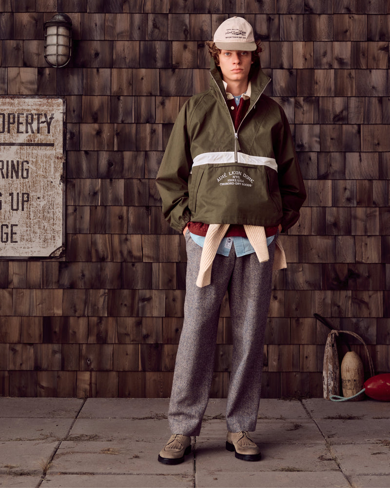 Person in an olive green jacket, gray pants, cap, and Suede Lug Sole Derby stands by a shingled wall with nautical props.