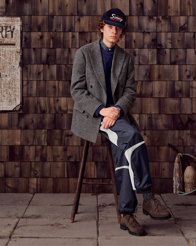 Young man wearing Suede Lug Sole Derby shoes, a blazer, cap, and sporty pants sits on a stool by a shingle wall.
