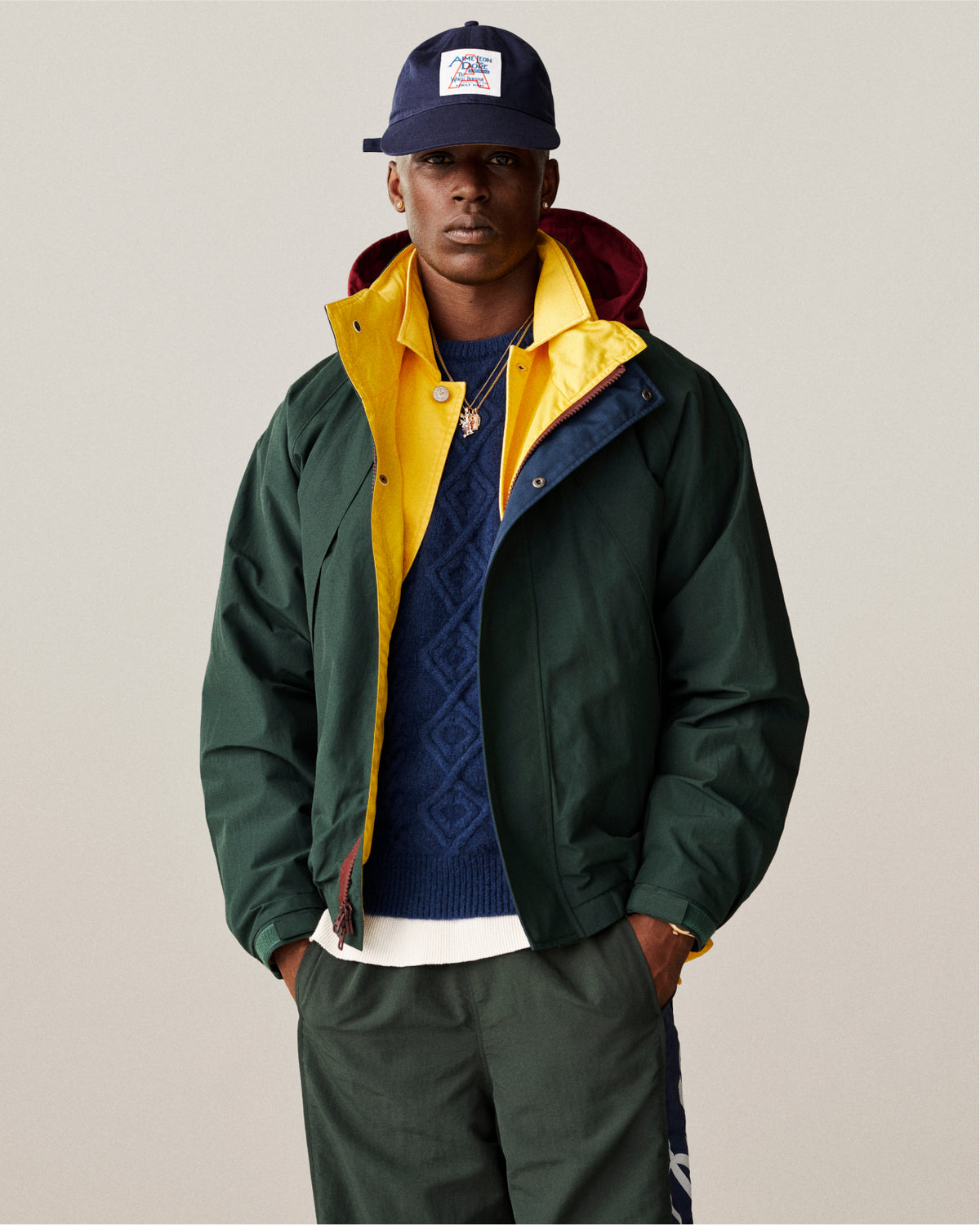 Man wearing a navy hat and green jacket, featuring the 14Kt Gold Musician Pendant, against a plain background.