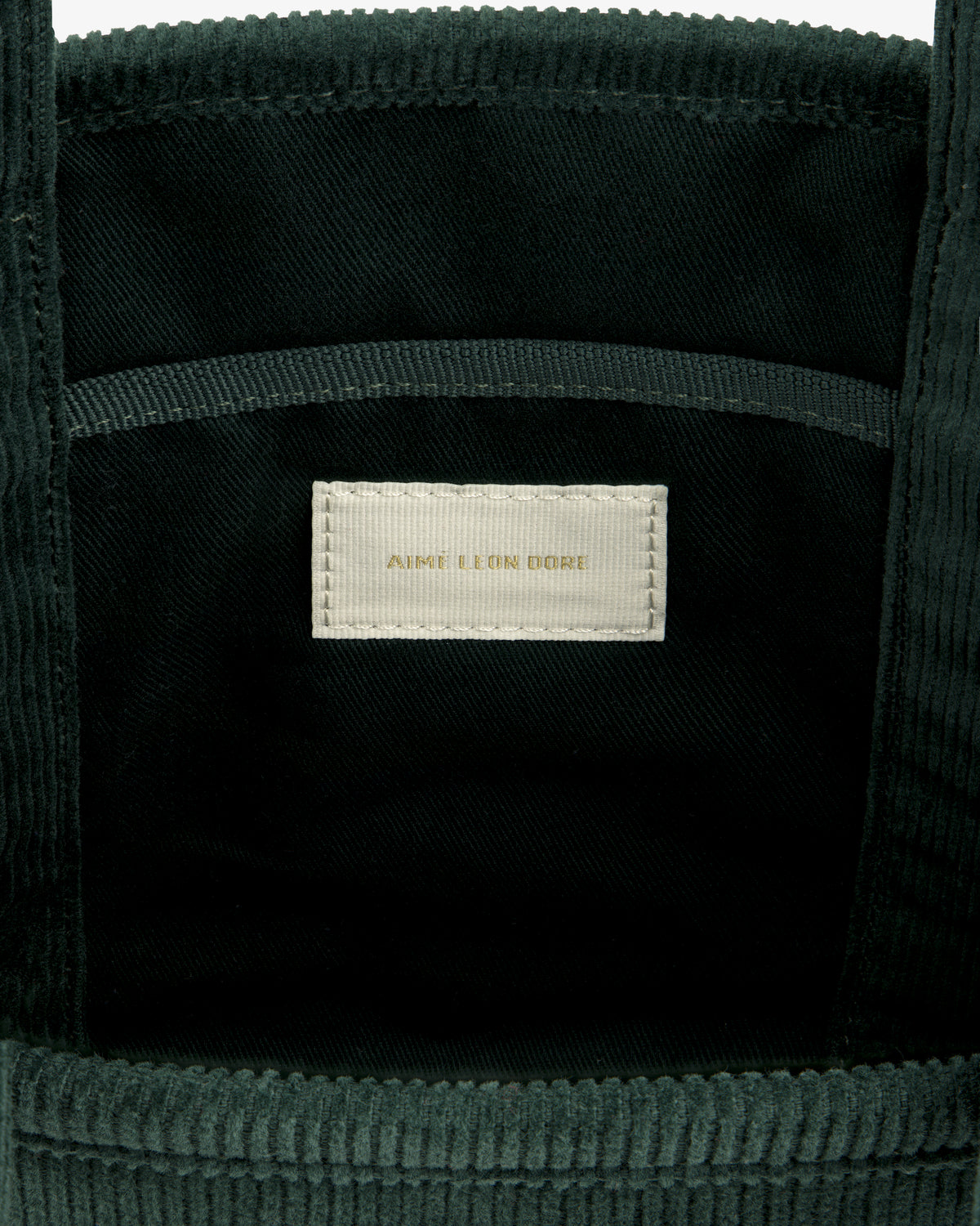 Interior of a dark green Mini Corduroy Unisphere Tote Bag featuring a central fabric label with AIME LEON DORE text.