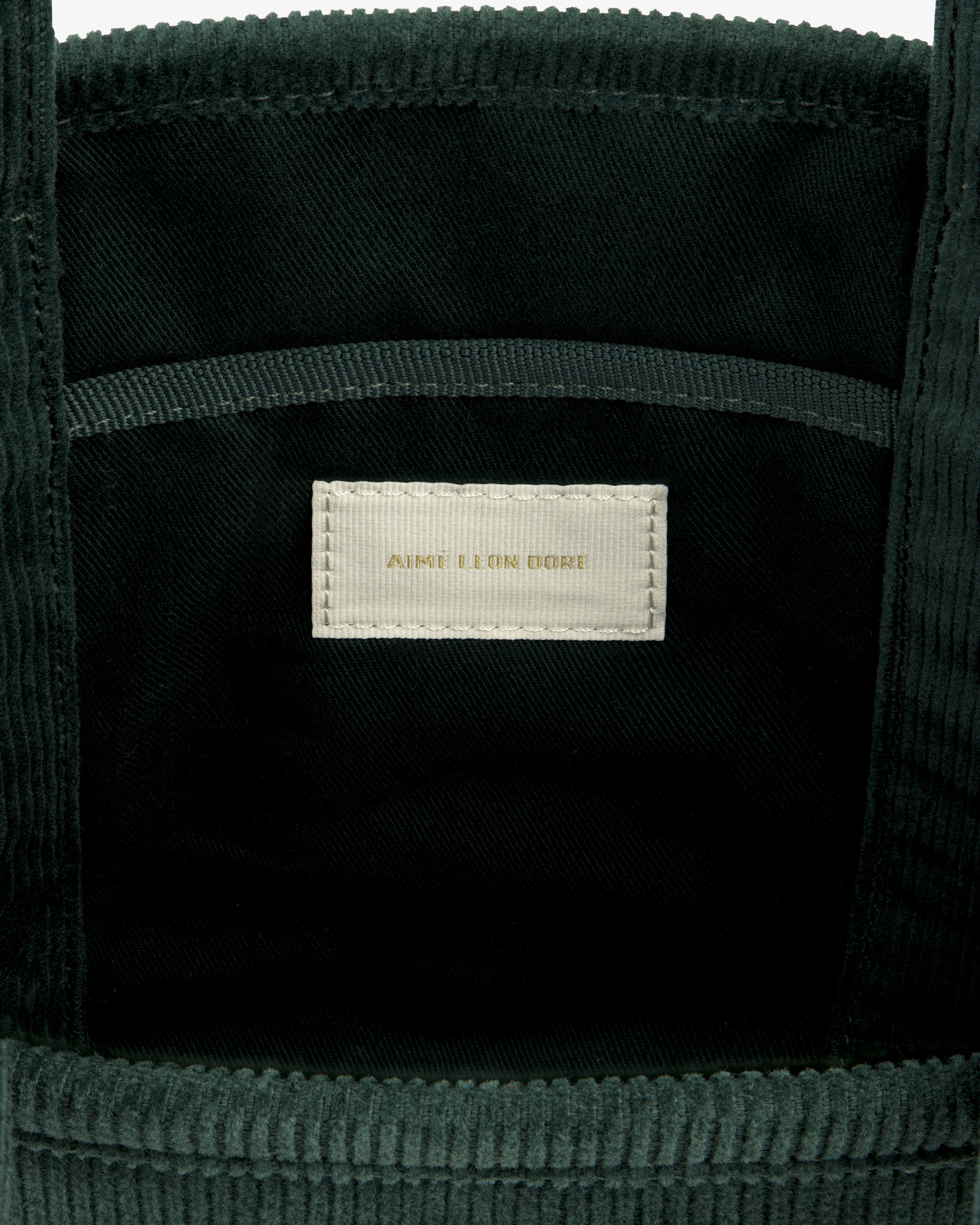 Interior of a dark green Mini Corduroy Unisphere Tote Bag featuring a central fabric label with AIME LEON DORE text.