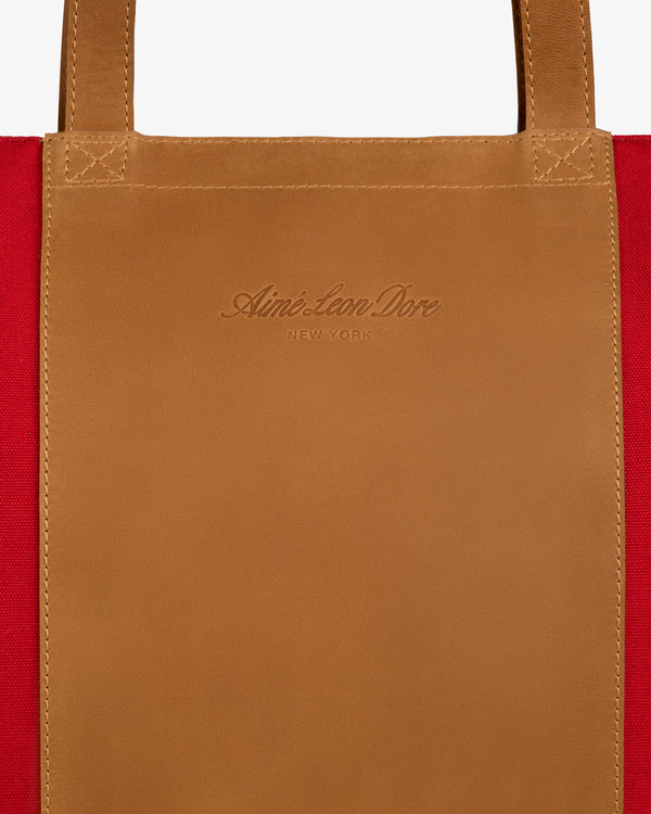 Close-up of Leather Canvas Zip Tote handle with Aimé Leon Dore New York embossed, red fabric background visible.