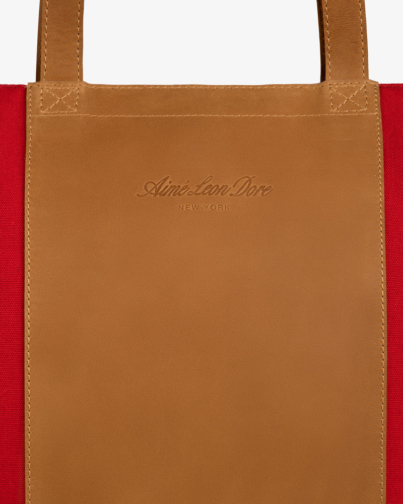 Close-up of Leather Canvas Zip Tote handle with Aimé Leon Dore New York embossed, red fabric background visible.
