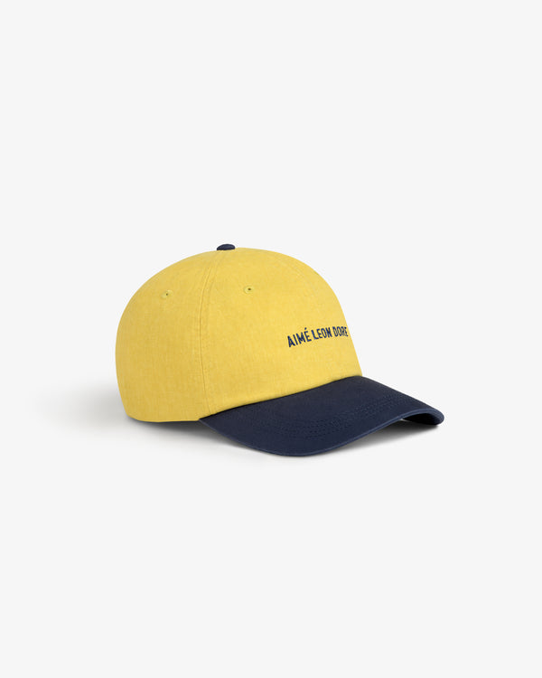 Pigment Dyed Logo Hat: yellow cap with navy visor and AIMÉ LEON DORE text, shown on white background.