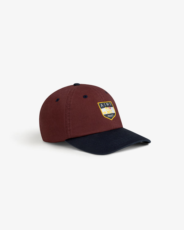 Crest Shield Hat: Burgundy cap with black visor, yellow/black shield patch front, and ventilation eyelets.