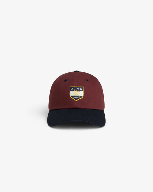 The Crest Shield Hat is burgundy with a navy brim and a yellow, blue, and white AIME crest on the front.