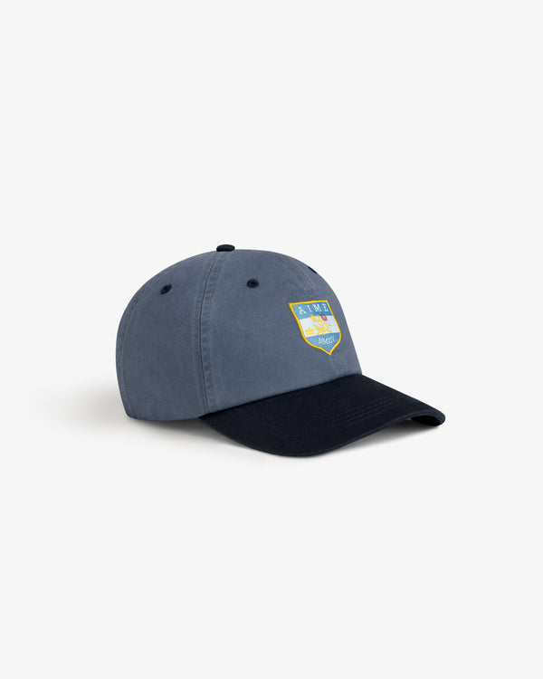 Crest Shield Hat in blue with a yellow and blue embroidered front patch, shown on a white background.