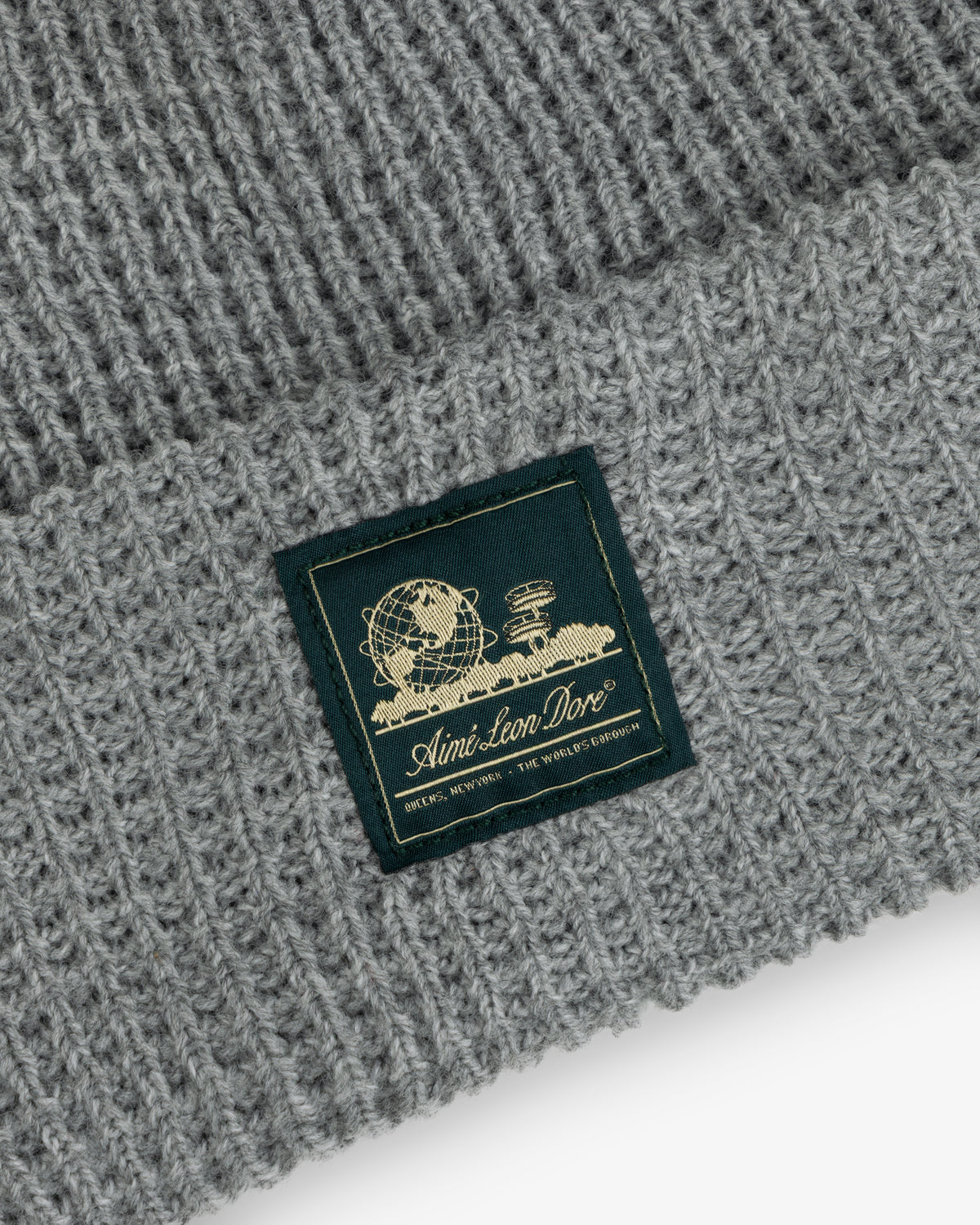 Close-up of a gray Waffle Knit Beanie with a patch showing a globe, books, and Aimé Leon Dore text.