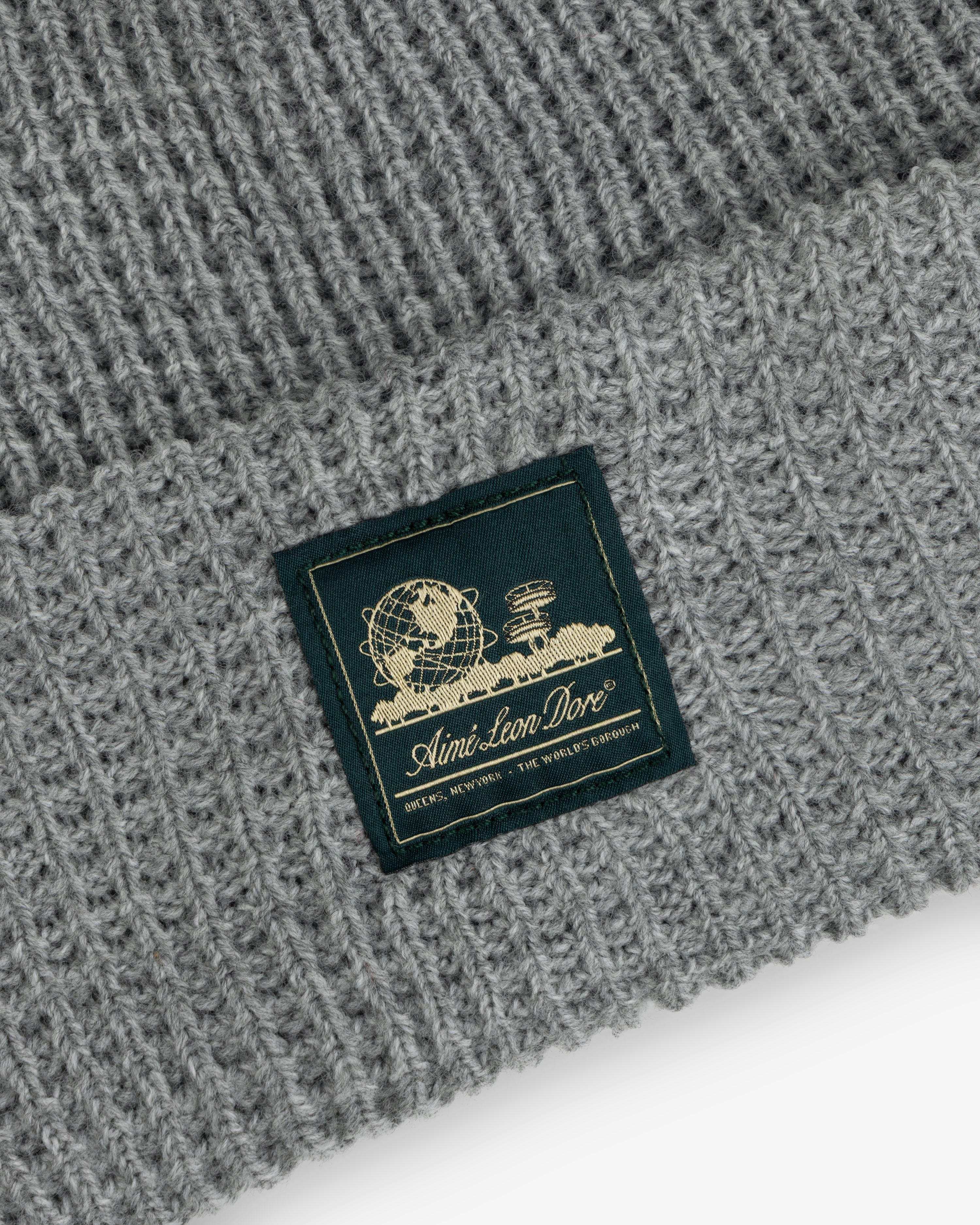 Close-up of a gray Waffle Knit Beanie with a patch showing a globe, books, and Aimé Leon Dore text.