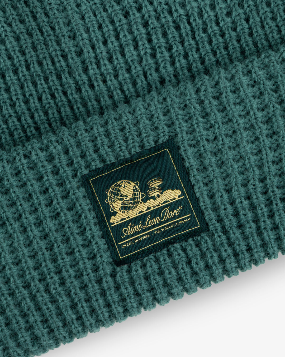 Close-up of a green Waffle Knit Beanie with a gold Aimé Leon Dore patch featuring a globe and books.