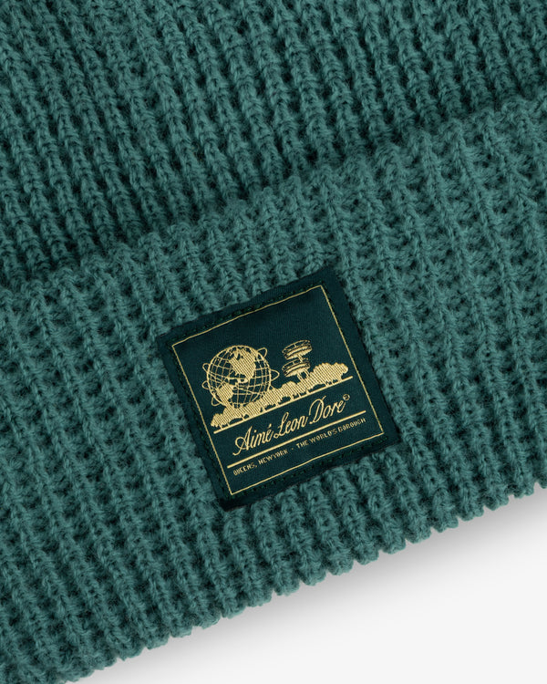 Close-up of a green Waffle Knit Beanie with a gold Aimé Leon Dore patch featuring a globe and books.
