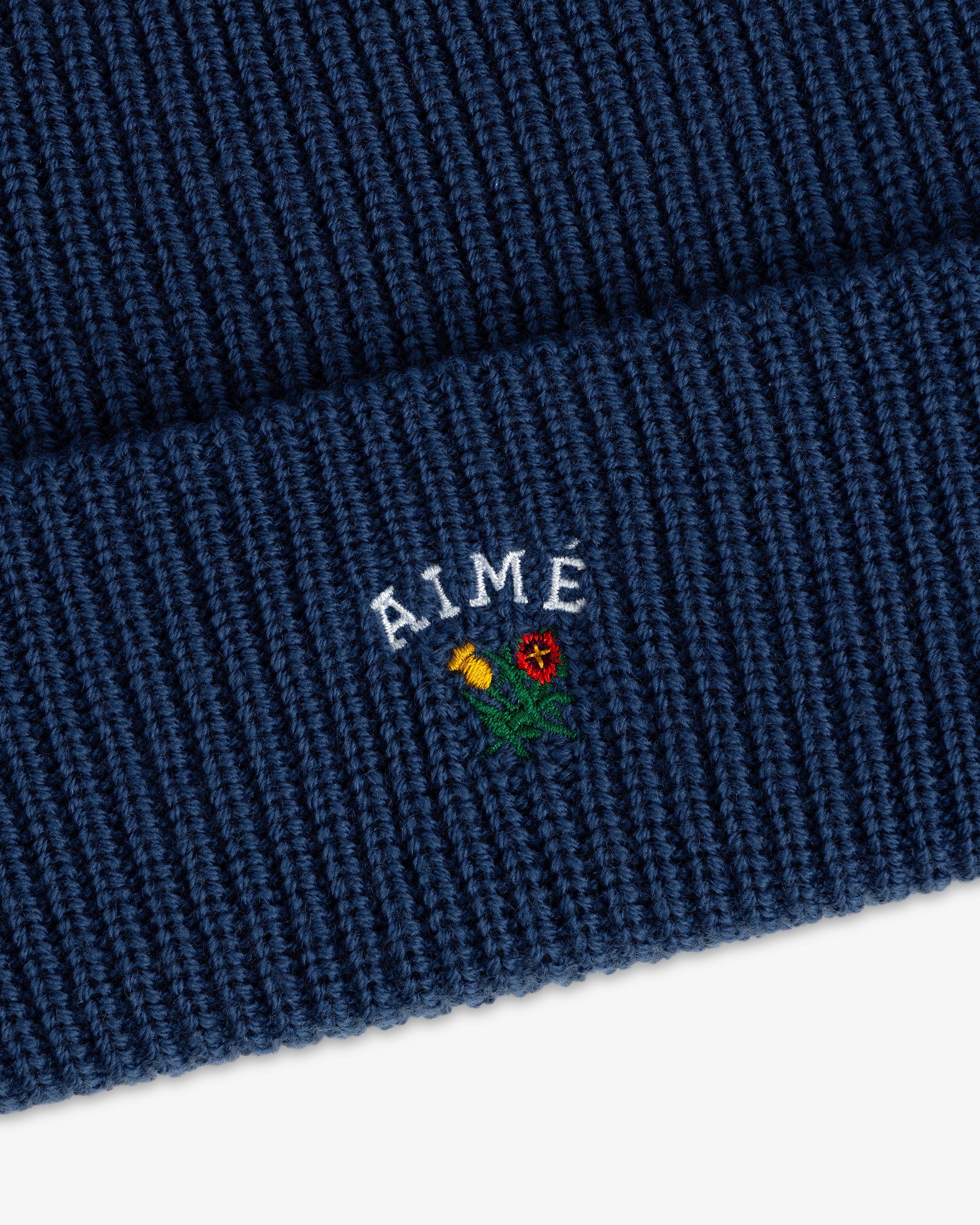 Blue Crest Merino Wool Beanie with AIME and a small yellow/red flower embroidered on the fold.