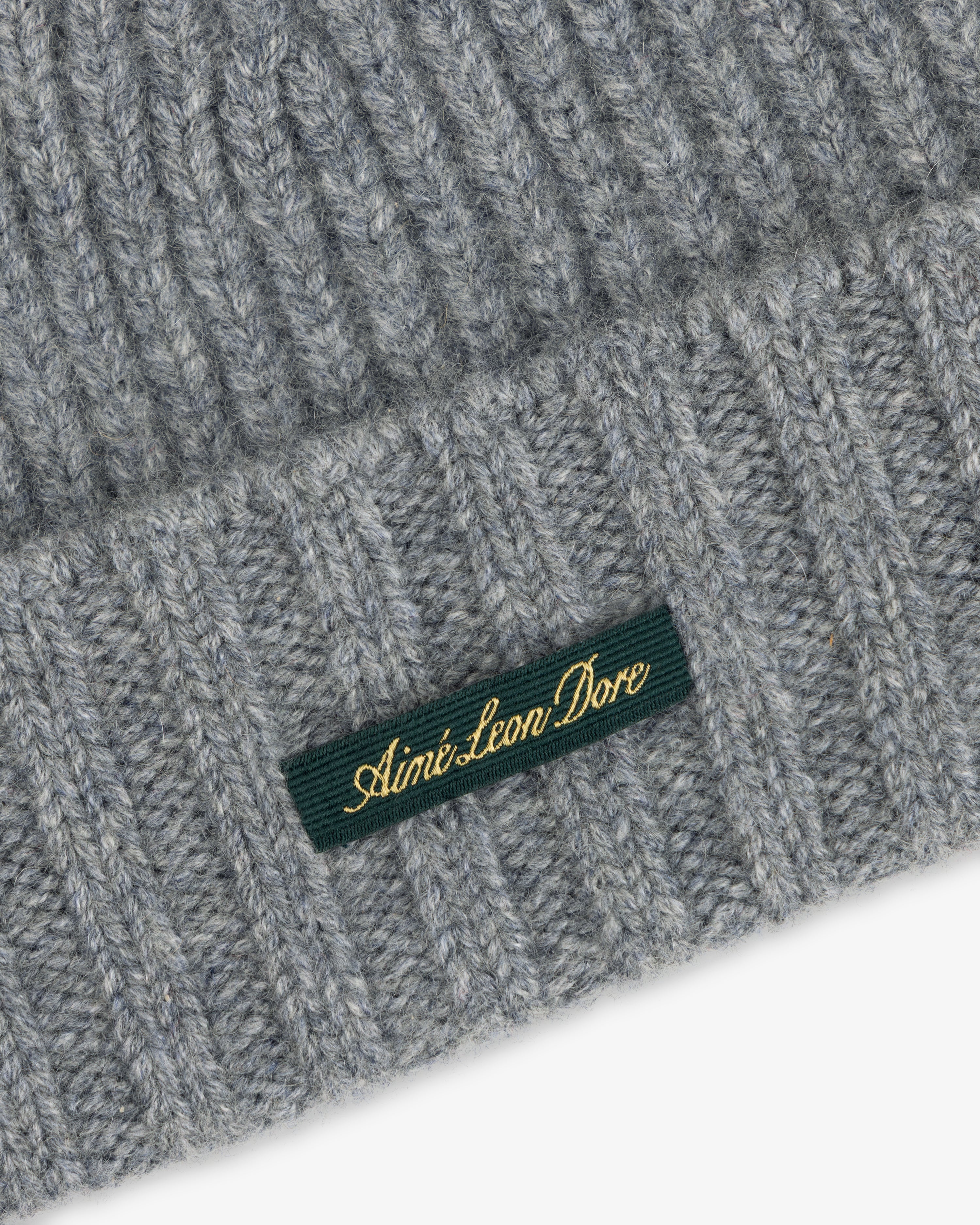 Close-up of a grey ribbed Cashmere Beanie with a green woven label featuring gold embroidered text.