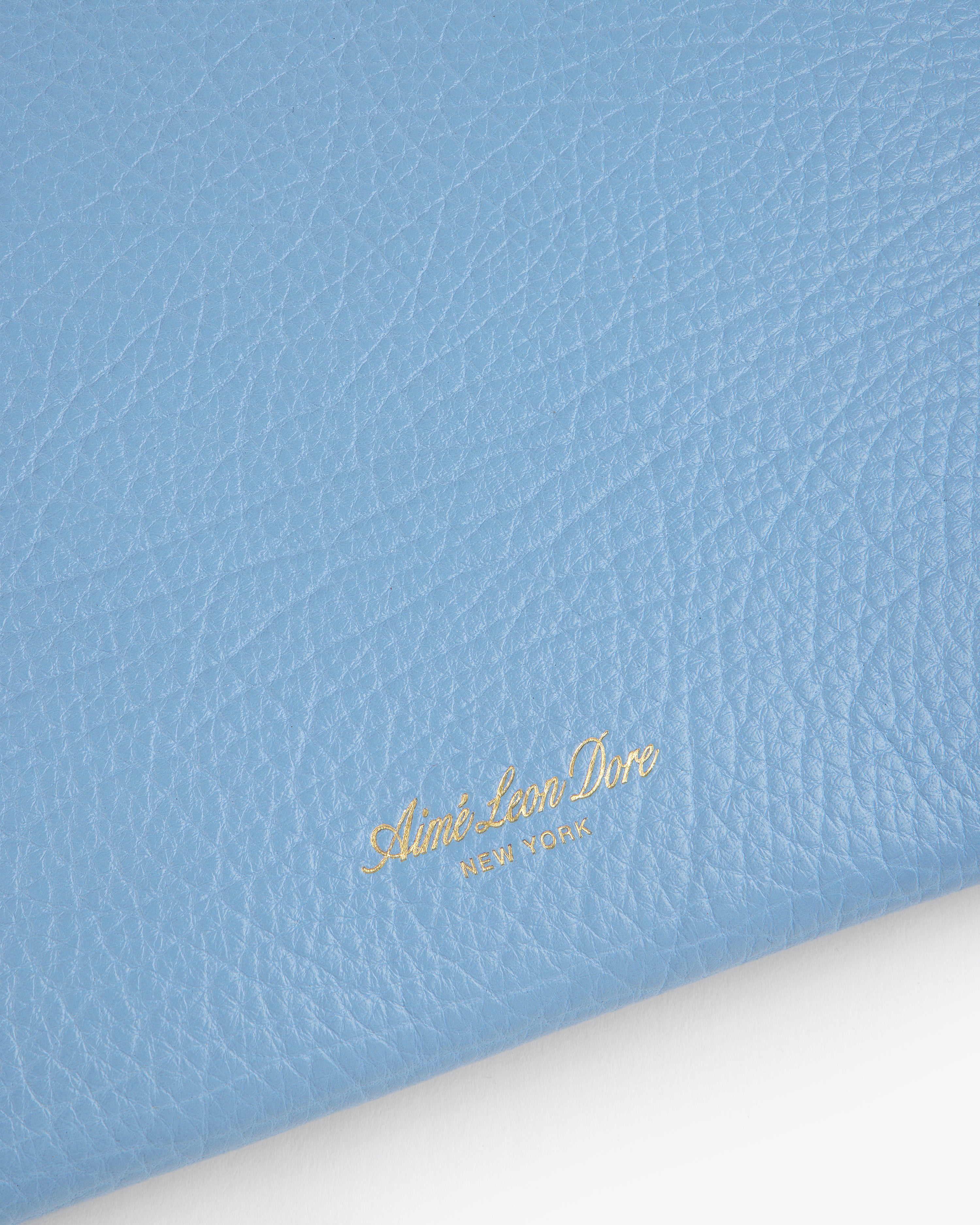 Close-up of textured blue Small Tumbled Leather Pouch with gold Aimé Leon Dore NEW YORK logo in lower left corner.