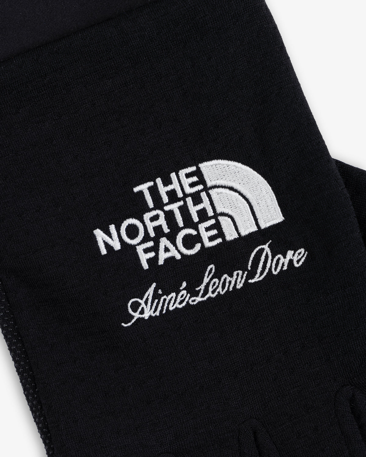 Close-up of ALD / The North Face DOTKNIT™ Liner Gloves with white embroidered logos on black fabric.
