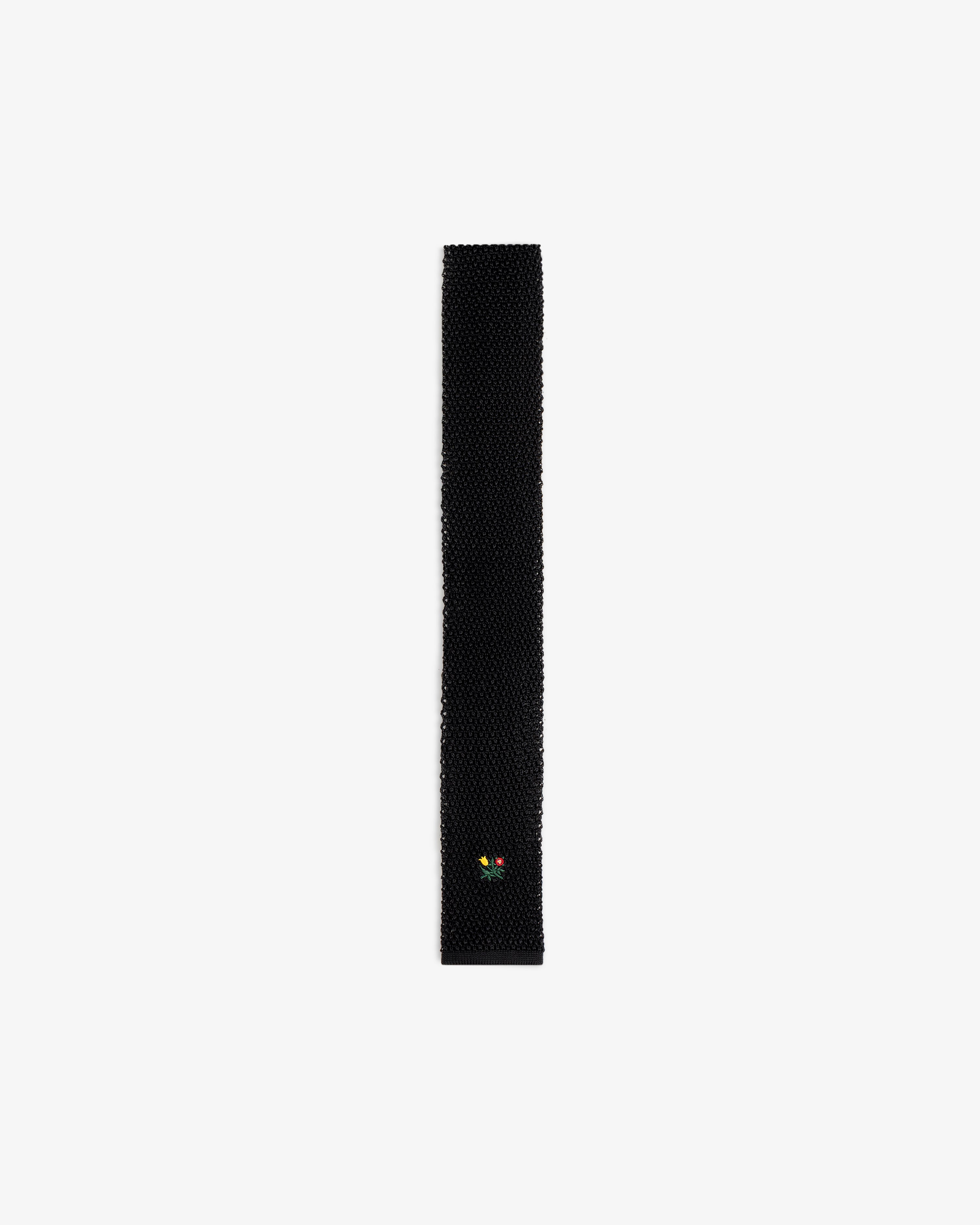 Crest Silk Knit Tie in black, featuring a small embroidered flower emblem near the tip.