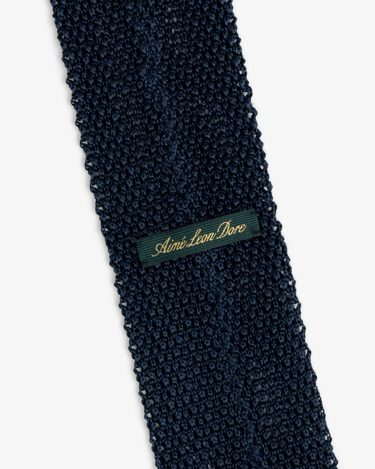 Dark blue Crest Silk Knit Tie with a rectangular tag reading Aimé Leon Dore in gold cursive.