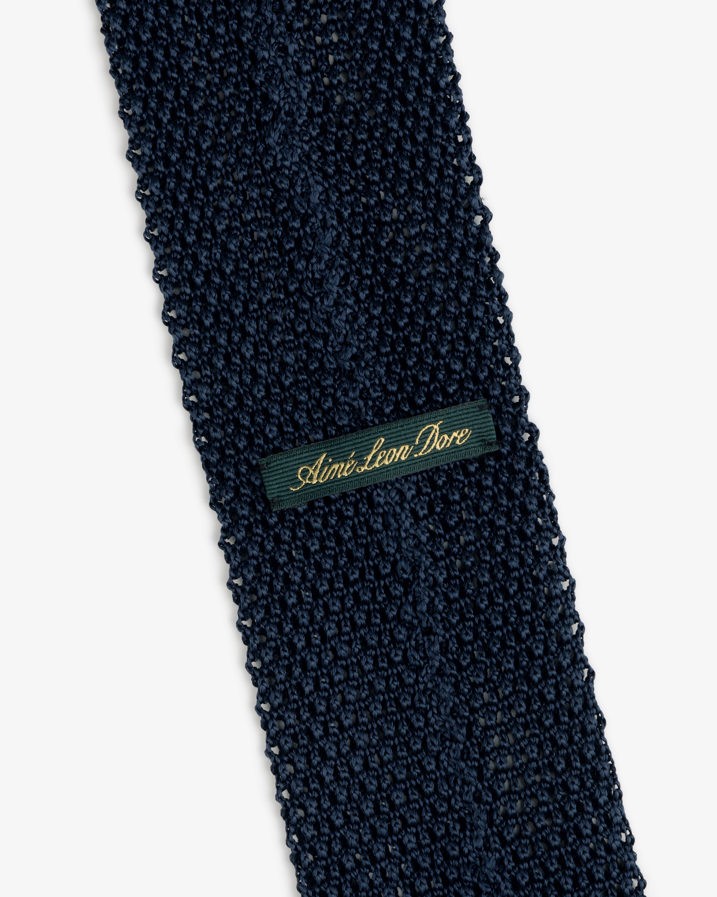 Dark blue Crest Silk Knit Tie with a rectangular tag reading Aimé Leon Dore in gold cursive.