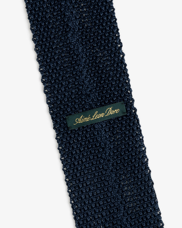 Dark blue Crest Silk Knit Tie with a rectangular tag reading Aimé Leon Dore in gold cursive.