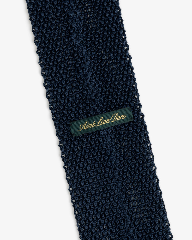 Dark blue Crest Silk Knit Tie with a rectangular tag reading Aimé Leon Dore in gold cursive.