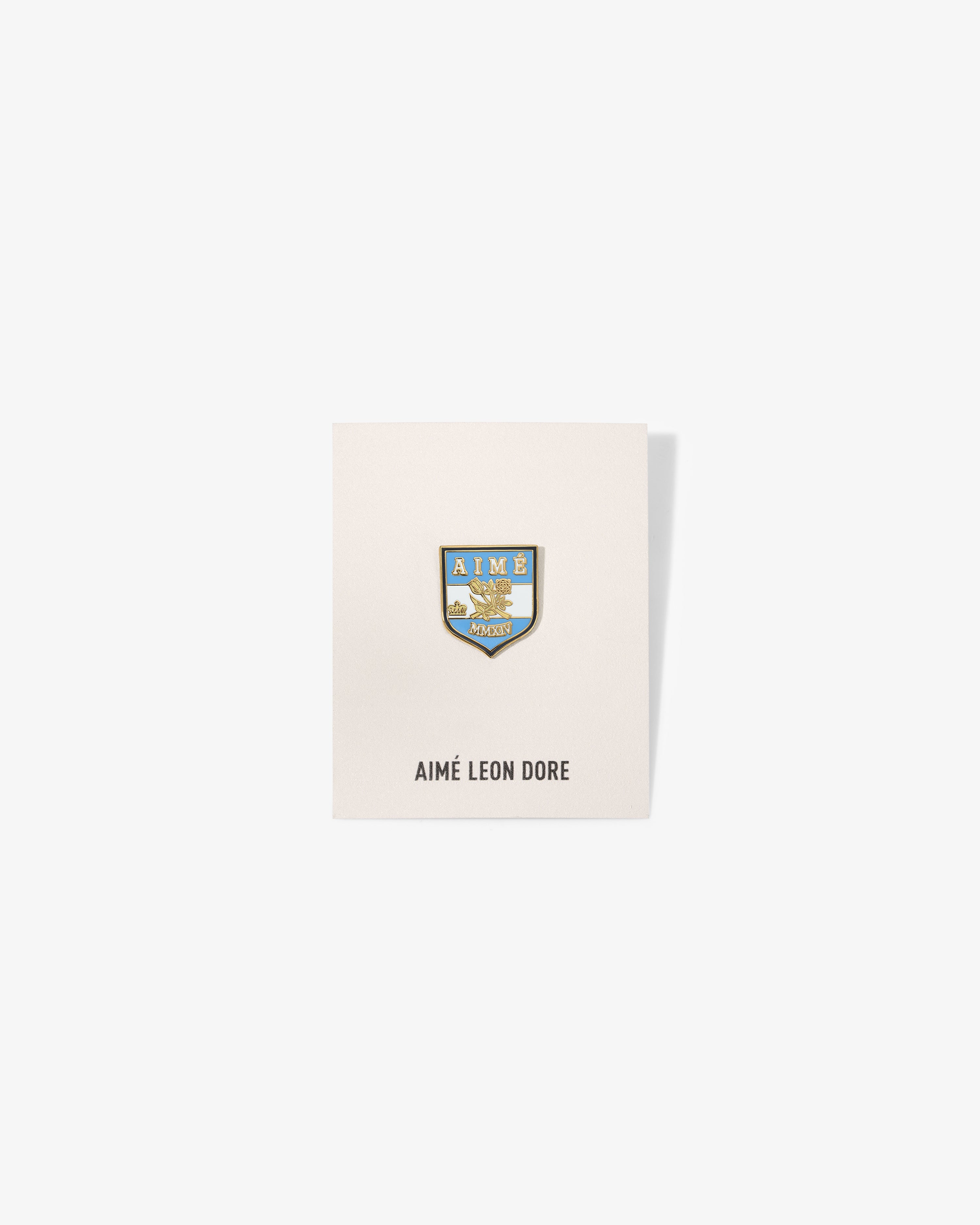 A Graphic Pin with blue and gold crest on a white card labeled AIMÉ LEON DORE below.