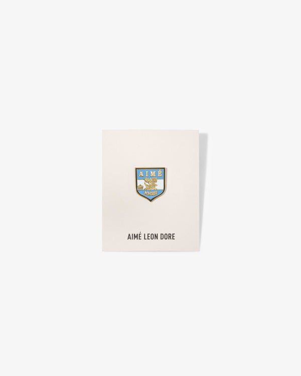 A Graphic Pin with blue and gold crest on a white card labeled AIMÉ LEON DORE below.