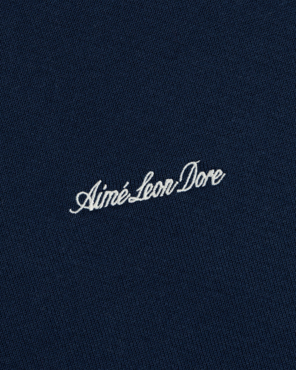 White cursive Aimé Leon Dore embroidery centered on the dark blue fabric of a Script Full Zip Hoodie.