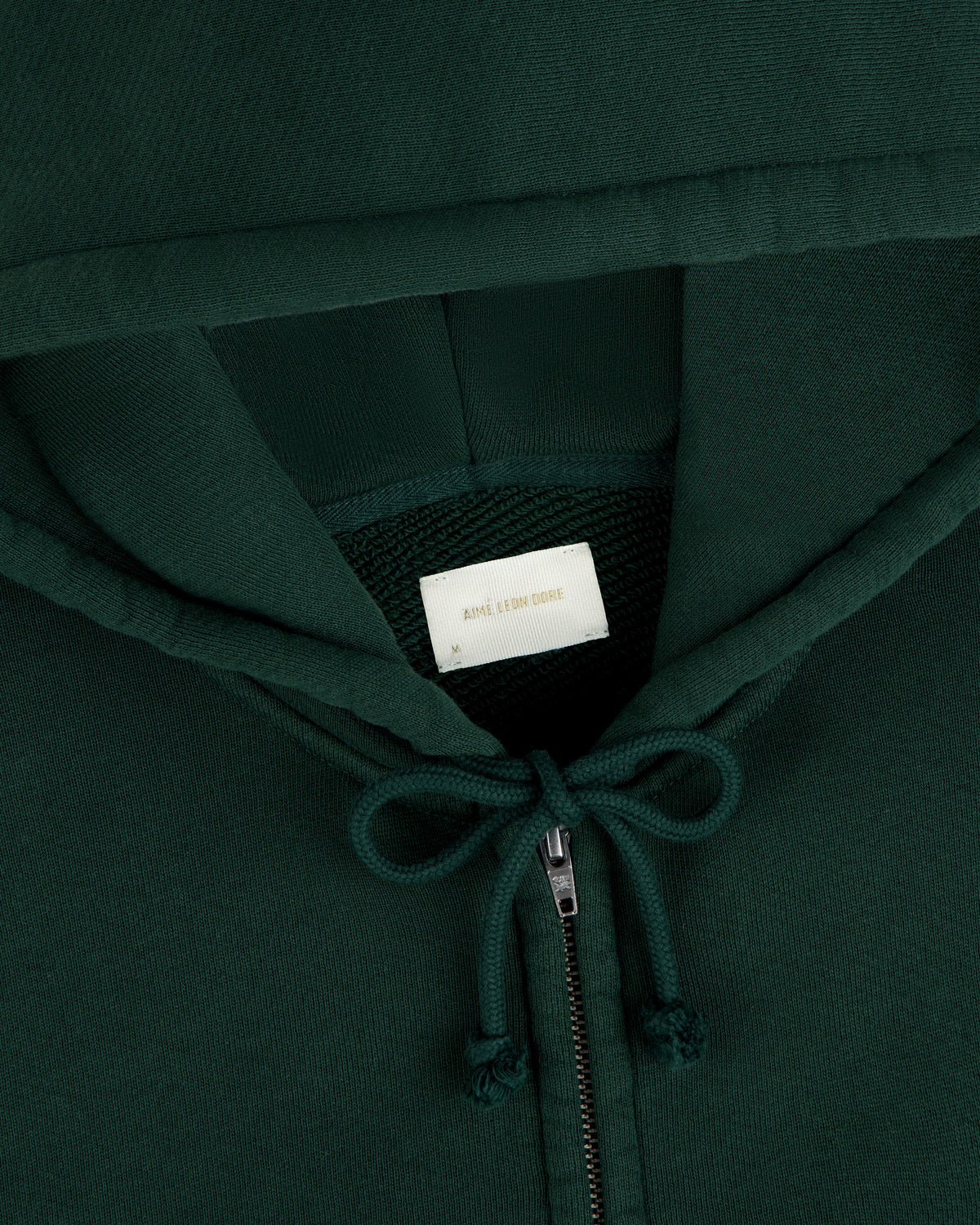 Close-up of a dark green Script Full Zip Hoodie with a white interior neck label, gold text, and a drawstring hood.
