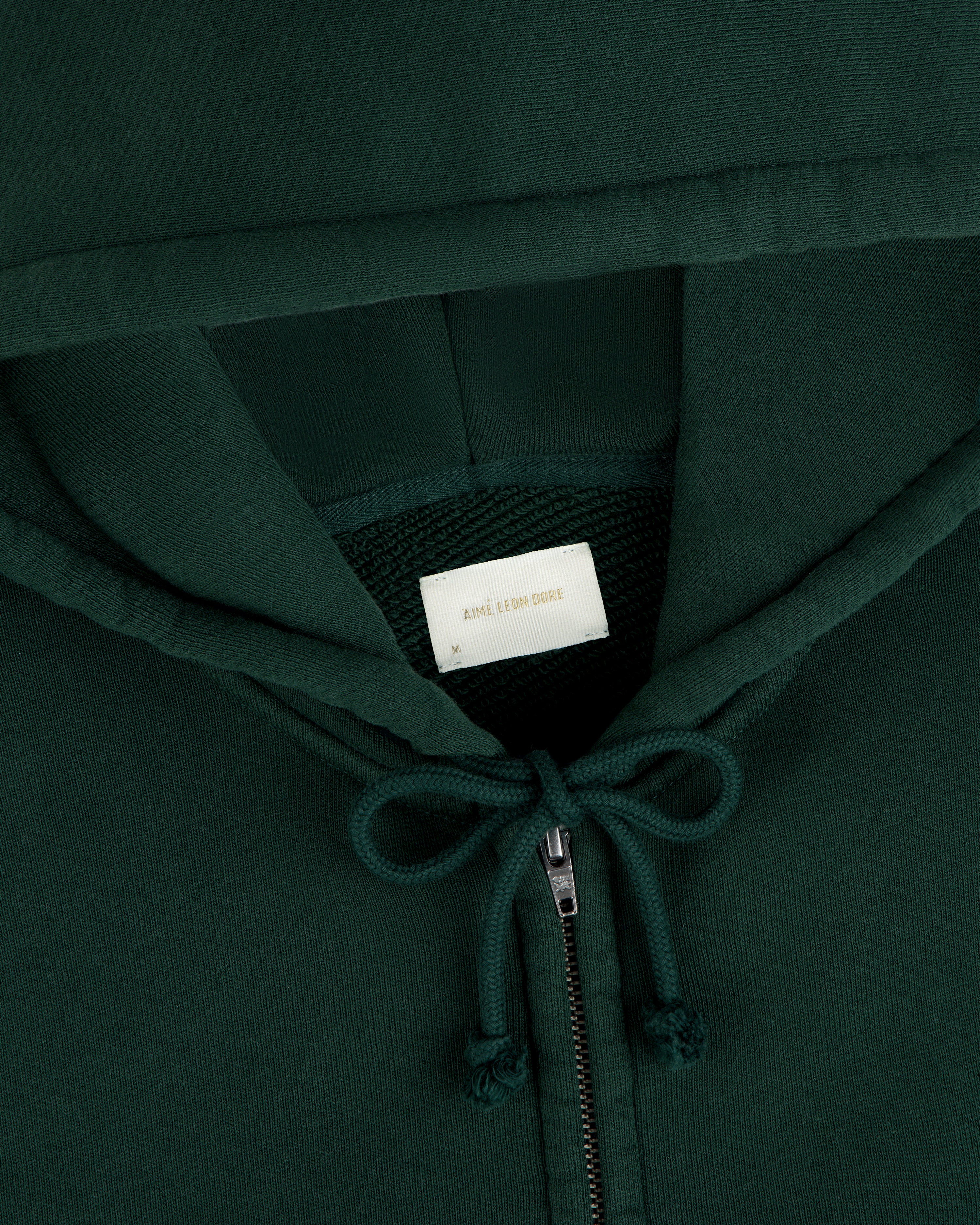 Close-up of a dark green Script Full Zip Hoodie with a white interior neck label, gold text, and a drawstring hood.