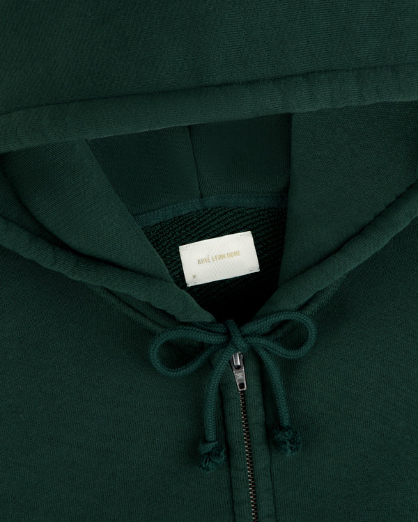 Close-up of a dark green Script Full Zip Hoodie with a white interior neck label, gold text, and a drawstring hood.