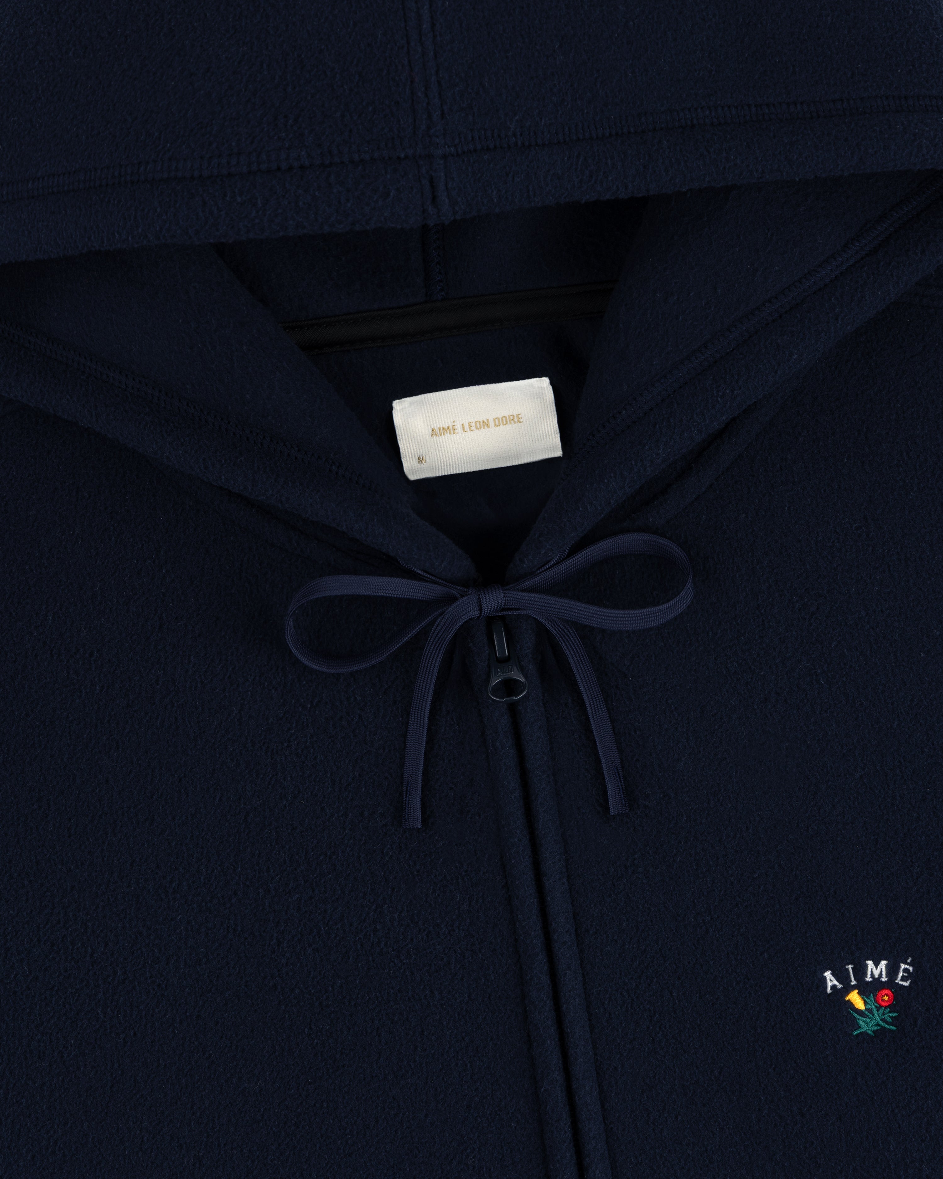 Detail of Crest Fleece Full-Zip Hoodie in navy with bow at neck and AIMÉ plus flower embroidered on chest.