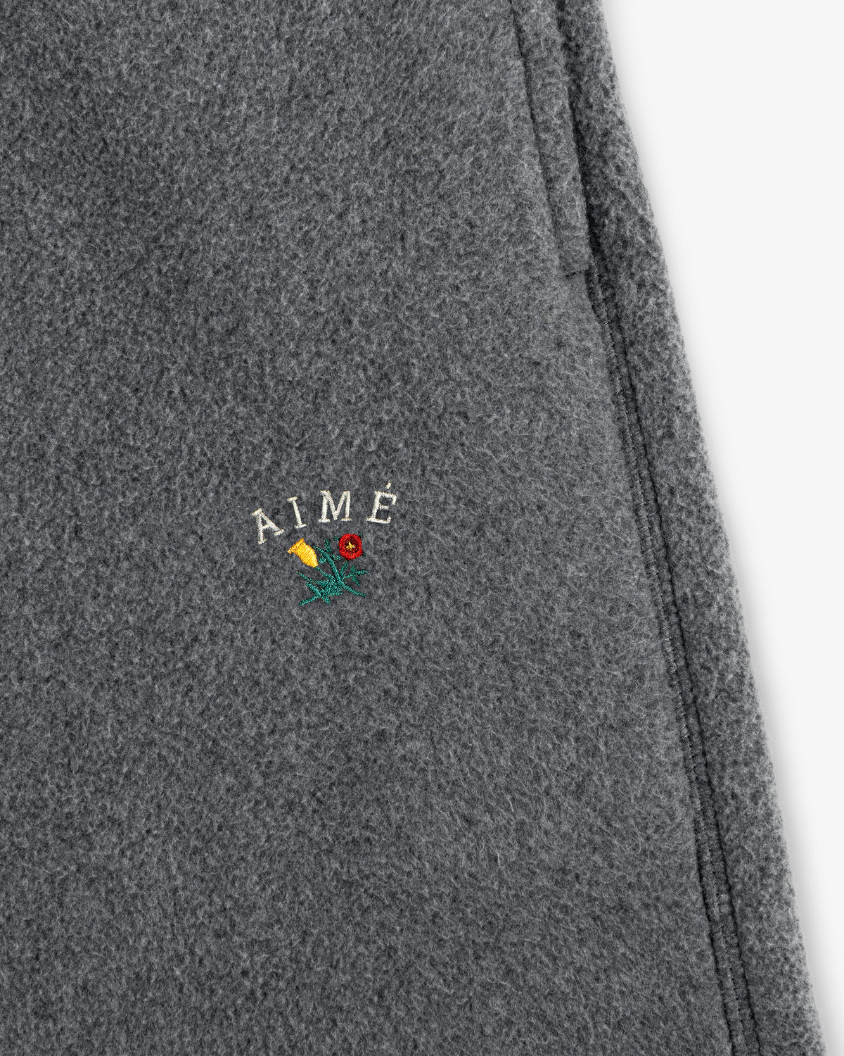 Close-up of gray Crest Fleece Sweatpant with AIMÉ and a small embroidered yellow and red flower detail.