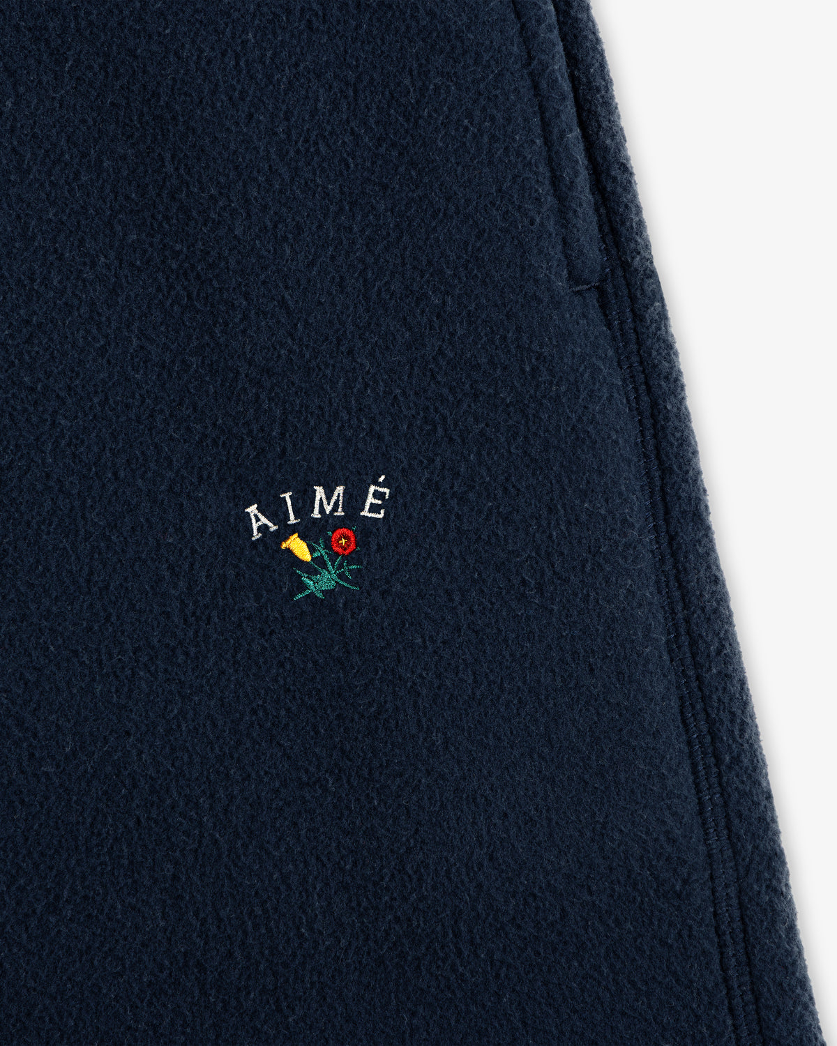 Close-up of Crest Fleece Sweatpant in navy with AIMÉ and small yellow and red embroidered flowers below the text.